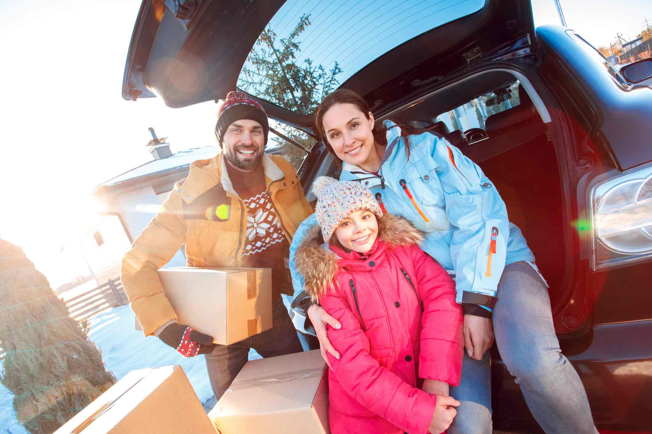 A family is bundled up in winter clothing and stands near the back of an open SUV, surrounded by moving boxes.