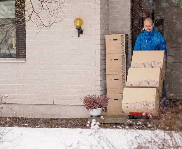 A man in a blue jacket moves several boxes in the snow from his home on a red dolly.