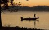 The silhouette of a man on a bass boat fishing at dawn on a lake.