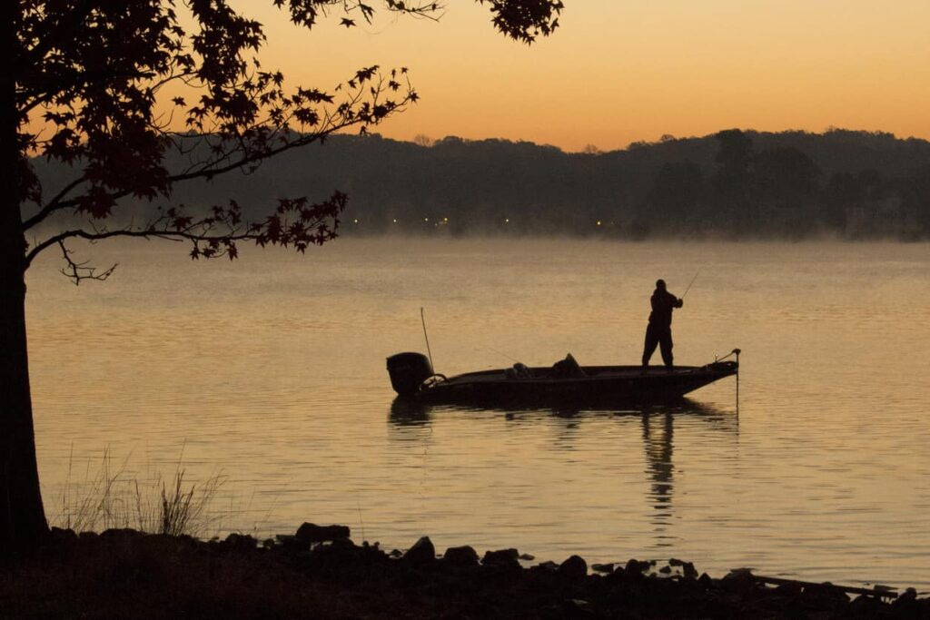 The silhouette of a man on a bass boat fishing at dawn on a lake.