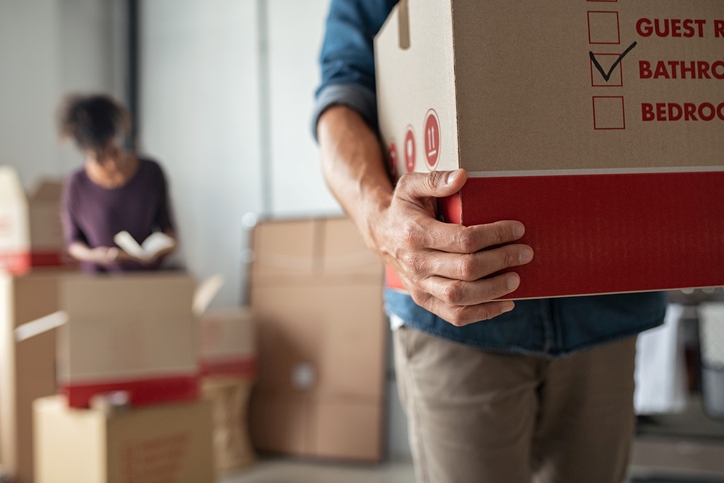 A man carries a cardboard box while moving with a woman in the background.