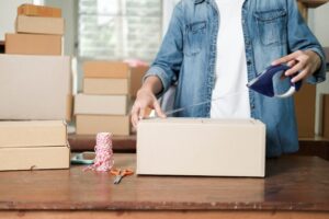A person in a chambray shirt tapes a cardboard box shut with a pair of orange scissors and a spool of twine beside.