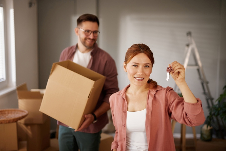 A happy couple smiles as the woman holds their keys and the man holds a cardboard box while moving.