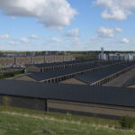 Large self-storage facility with rows of dark-roofed units, industrial buildings, and a water tower under blue sky.