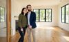 A young couple stands in a sunlit empty living room doorway, gazing around with hopeful expressions as homebuyers.