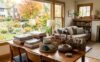 Sunlit dining table displays folded linens, ceramic dishes, and wooden keepsakes beside a window with autumn trees.